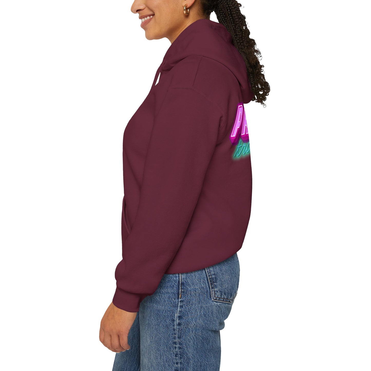 A person in a Protect Trans Kids Hoodie stands sideways, smiling. The maroon unisex hoodie features colorful text on the back and is made from ethically grown cotton, perfectly pairing with their blue jeans. Their curly hair is tied back against a plain white background.