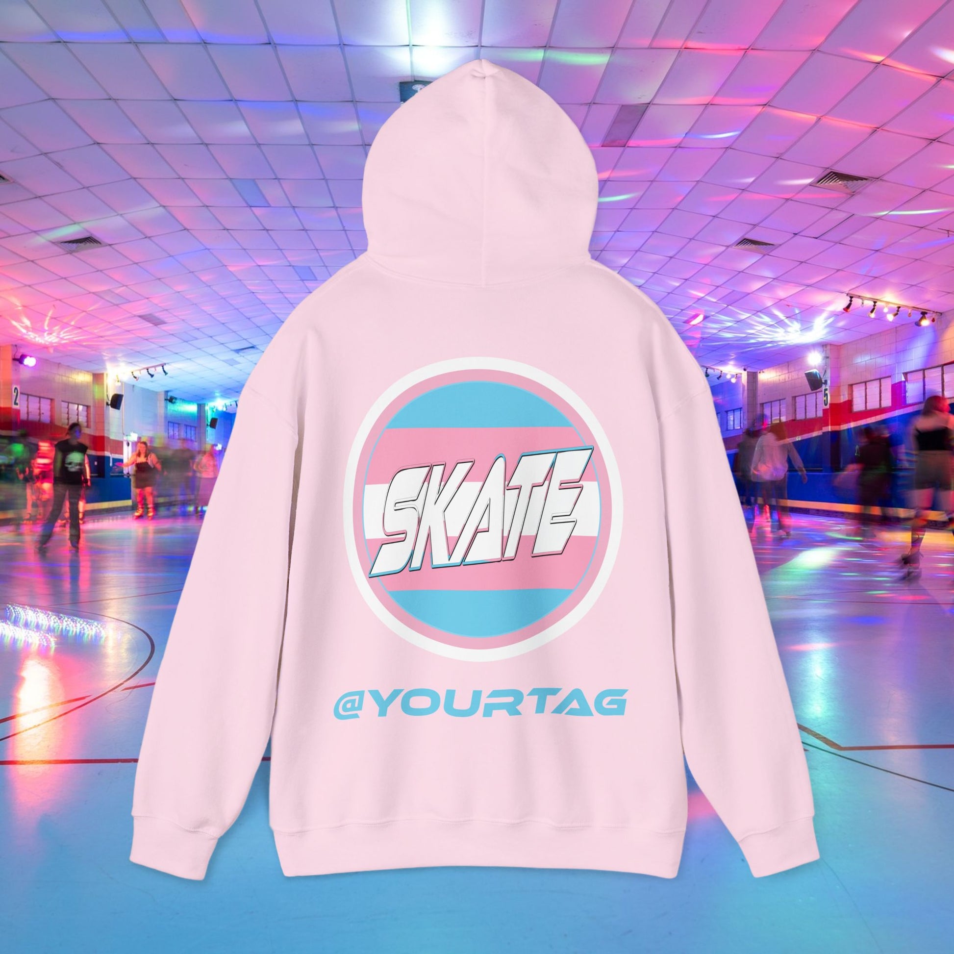 A person wearing a Printify "Add Custom Name - SKATE Trans Flag round logo Hoodie - Australian Shipping," a black unisex heavy blend hooded sweatshirt featuring the word "SKATE" inside a trans circle logo on the back with their skater name written beneath it. The background shows an indoor roller skating rink with colorful lights and other skaters.