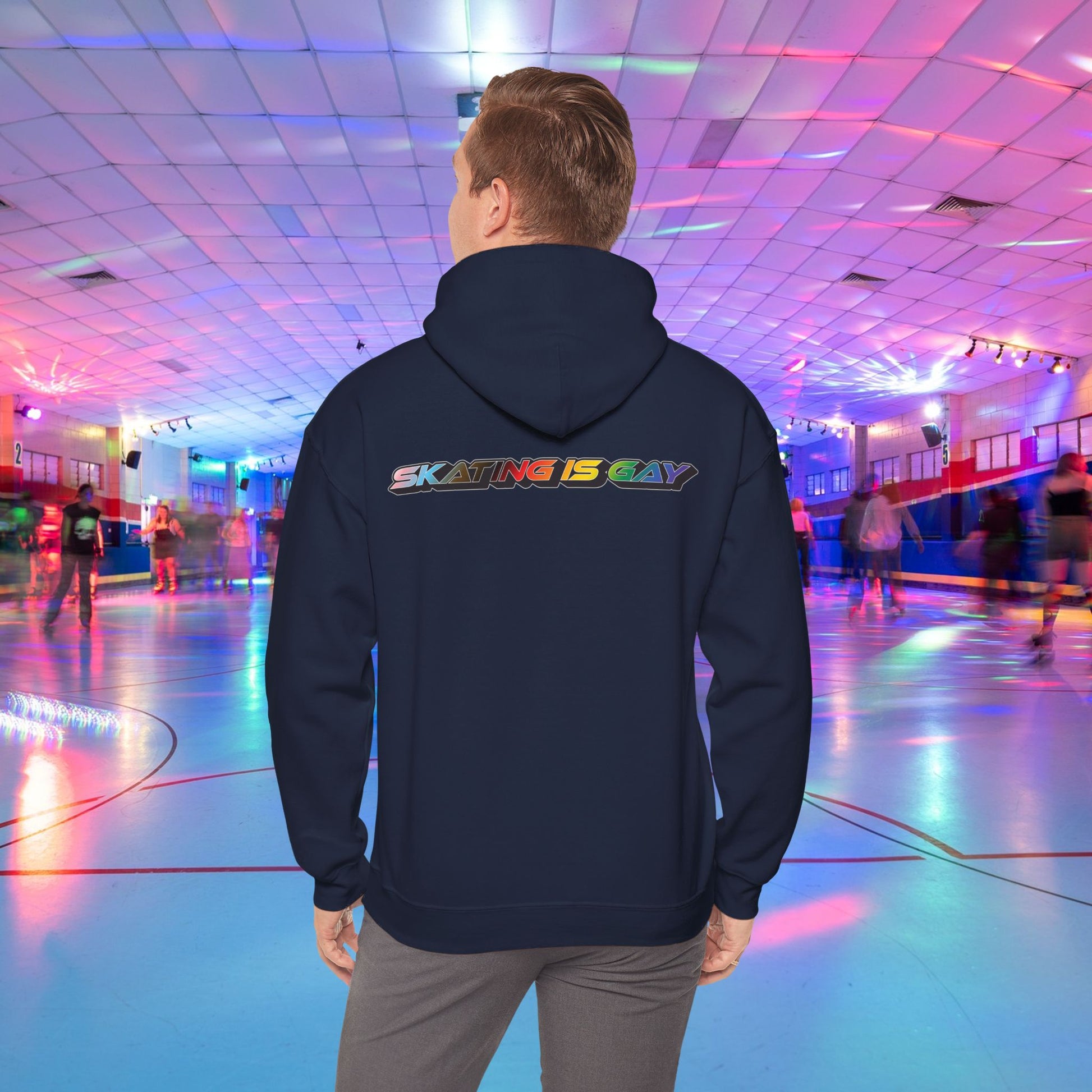 A person wearing a high-quality Skating Is Gay Hoodie by Printify, featuring the vibrant rainbow pride text "SKATING IS GAY" printed on the back, stands in the foreground of a neon-lit roller skating rink. People skate in the blurred background as colorful lights reflect off the rink's surface. The product is available with Australian shipping.