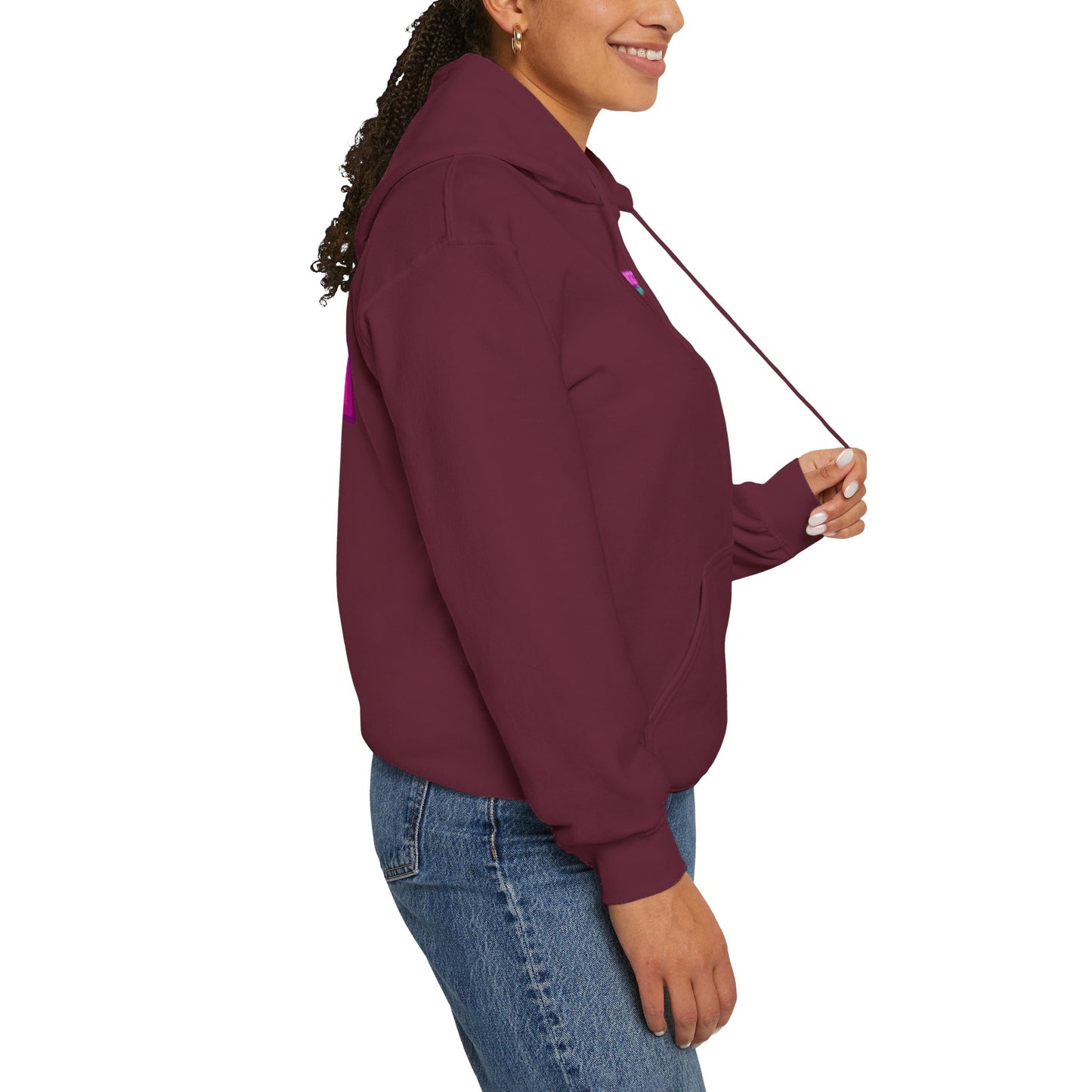 A person in a dark maroon Protect Trans Kids unisex hoodie made from ethically grown cotton and blue jeans smiles against a white background, holding one hoodie string with their right hand. Their hair is styled in a braid.