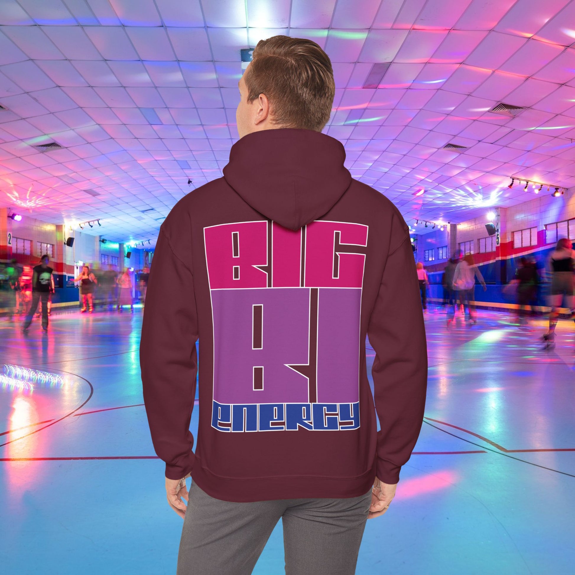 A person with light brown hair is wearing a high-quality BIG BI ENERGY Hoodie from GAY SKATE, featuring bold, Bisexual Pride Flag colour letters on the back. They are standing in a roller skating rink illuminated with vibrant, colorful lights. The cozy feel and warmth of this trans and queer-owned brand is evident.