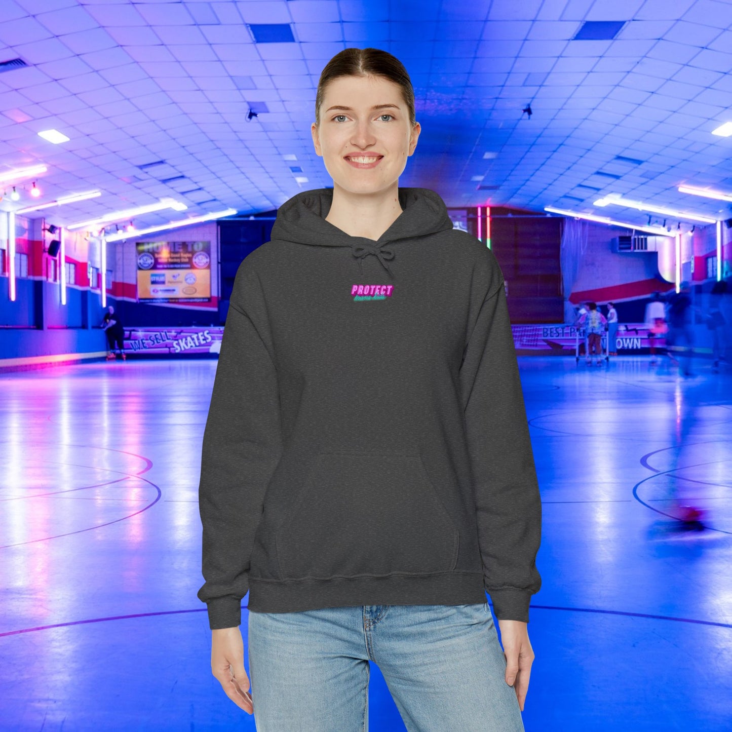 At a neon-lit skating rink, an individual wearing a "Protect Trans Kids Hoodie - Australian Shipping," crafted from ethically grown cotton, smiles as vibrant pink and blue lights illuminate blurred skaters in the background.