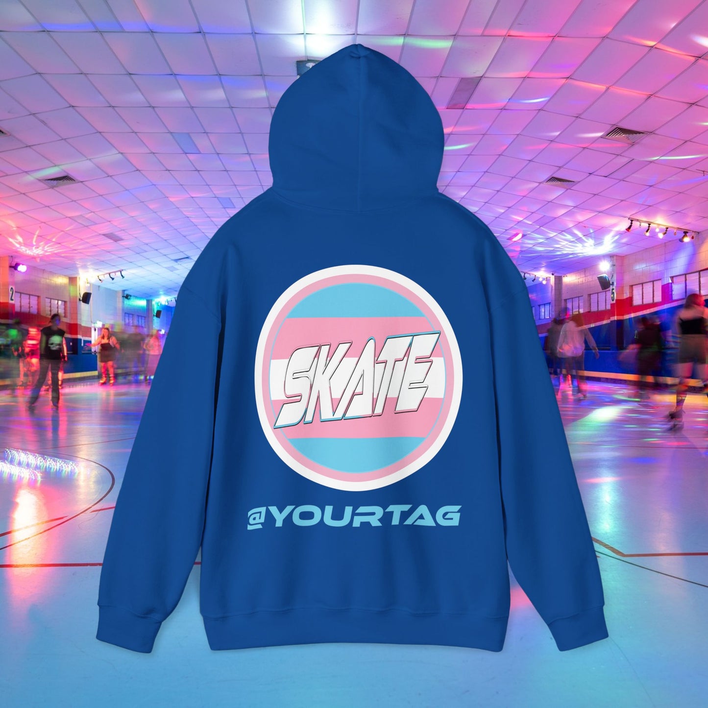 A person wearing a Printify "Add Custom Name - SKATE Trans Flag round logo Hoodie - Australian Shipping," a black unisex heavy blend hooded sweatshirt featuring the word "SKATE" inside a trans circle logo on the back with their skater name written beneath it. The background shows an indoor roller skating rink with colorful lights and other skaters.