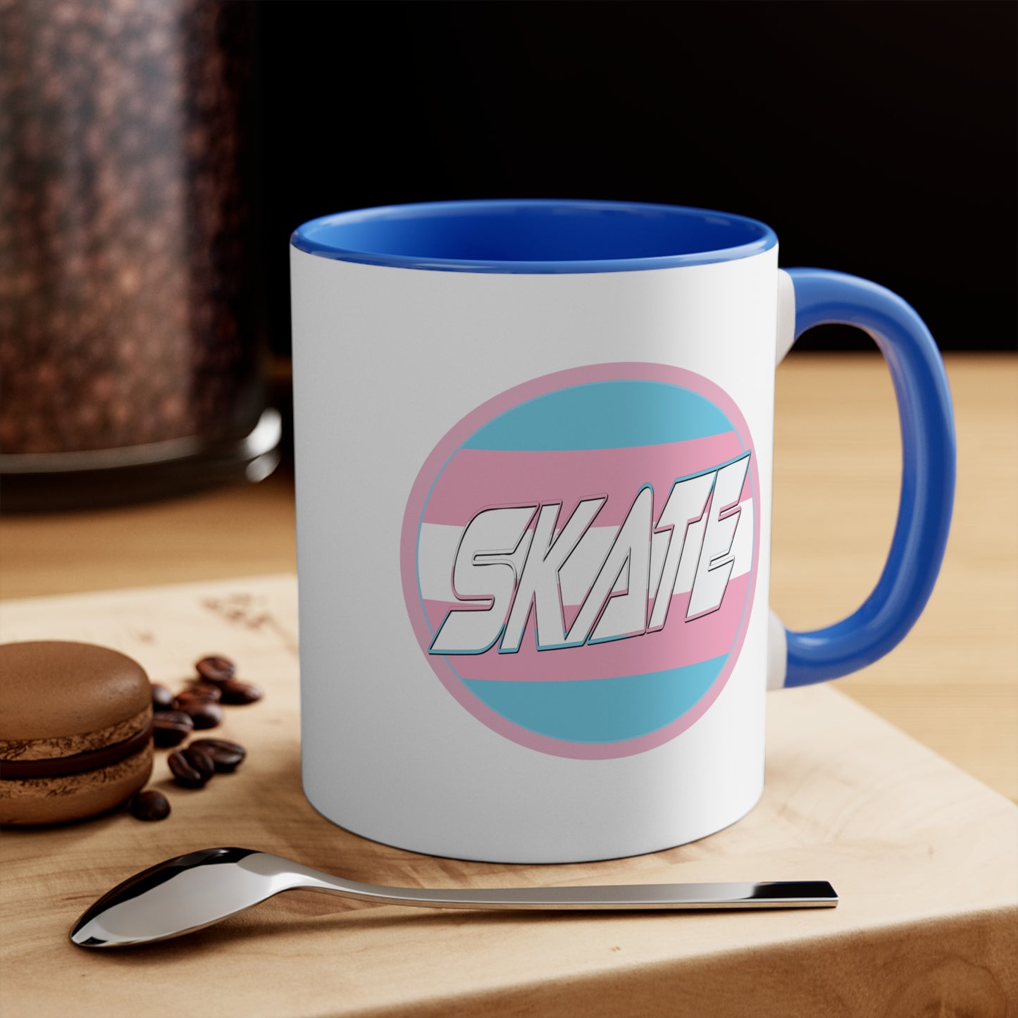 The SKATE trans flag circle design accent mug by Printify features a white exterior, blue handle, and blue interior. It showcases a circular design inspired by the trans flag, incorporating pink, light blue, and white colors. The word "SKATE" is prominently displayed in dynamic white letters at the center of the design.