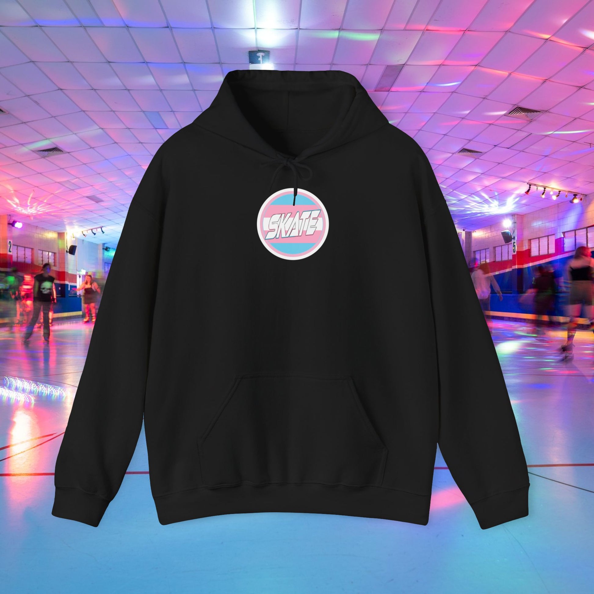 A person wearing a Printify "Add Custom Name - SKATE Trans Flag round logo Hoodie - Australian Shipping," a black unisex heavy blend hooded sweatshirt featuring the word "SKATE" inside a trans circle logo on the back with their skater name written beneath it. The background shows an indoor roller skating rink with colorful lights and other skaters.