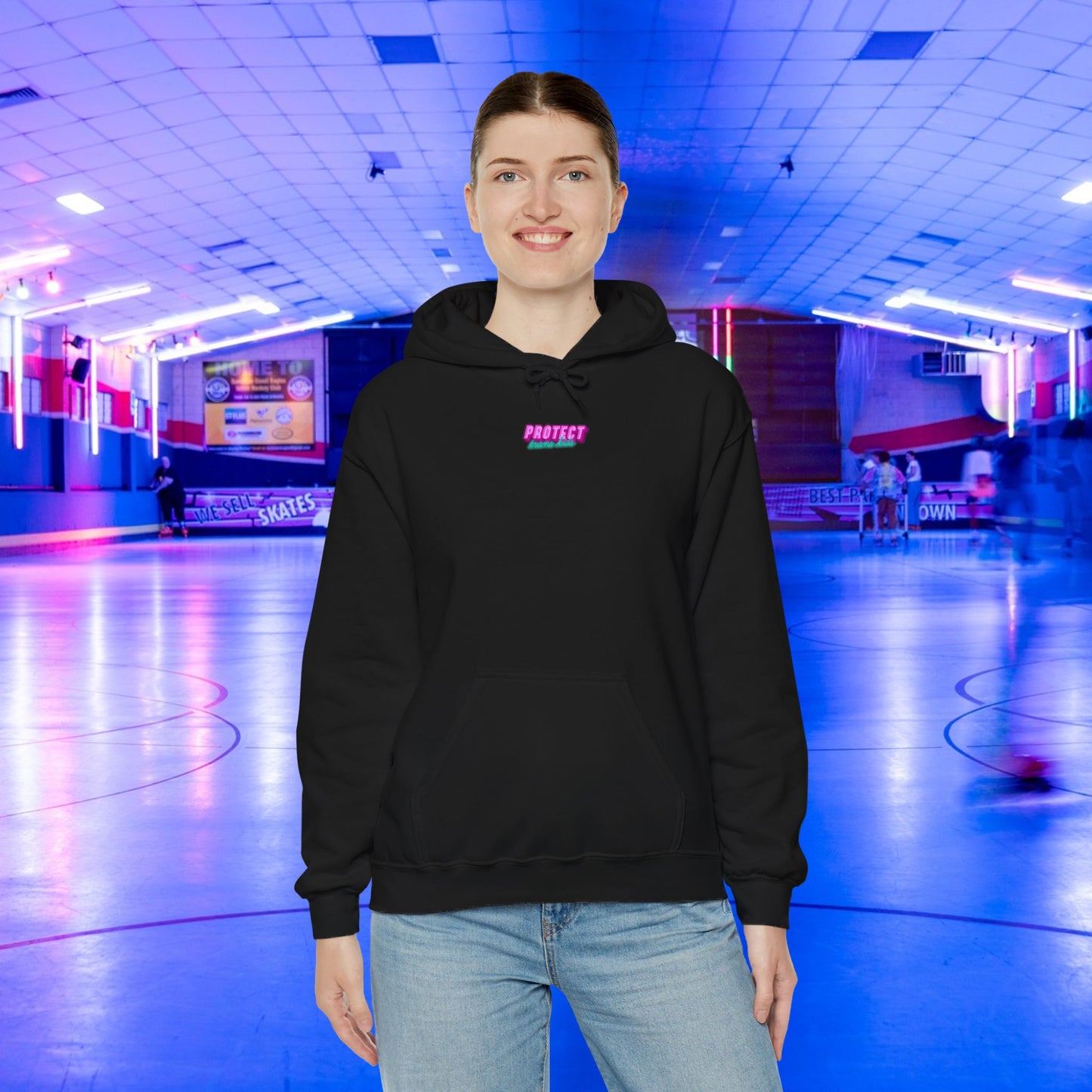 A person wearing a Protect Trans Kids Hoodie smiles in a neon-lit roller skating rink, glowing with blue and pink lights. In the background, people are joyfully skating.