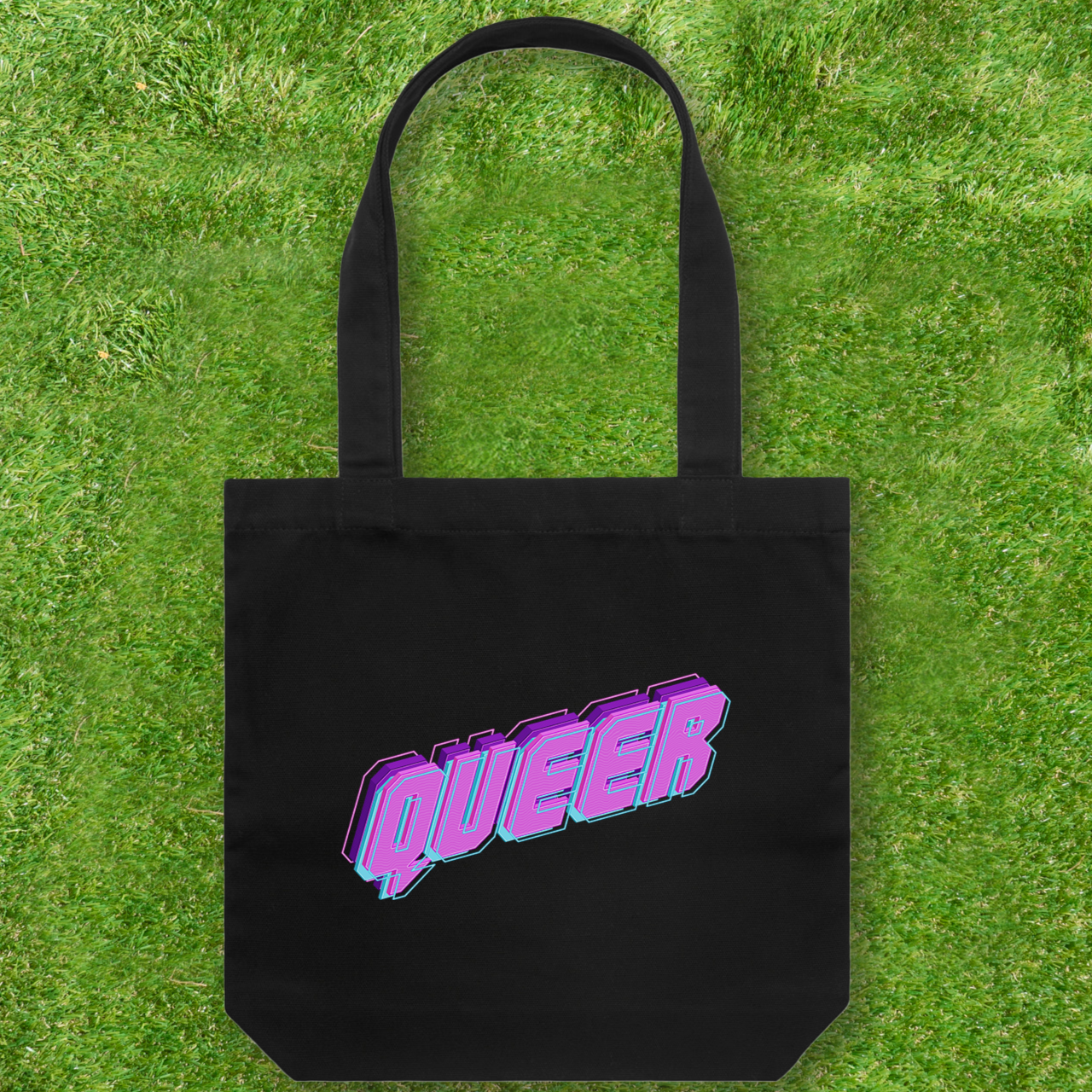 A large printed cotton canvas tote bag in black, featuring the word "QUEER" in bold, vibrant pink and blue retro-style letters, is elegantly placed on lush green grass. This stylish accessory from Gay Skate proudly supports queer and trans-owned brands.