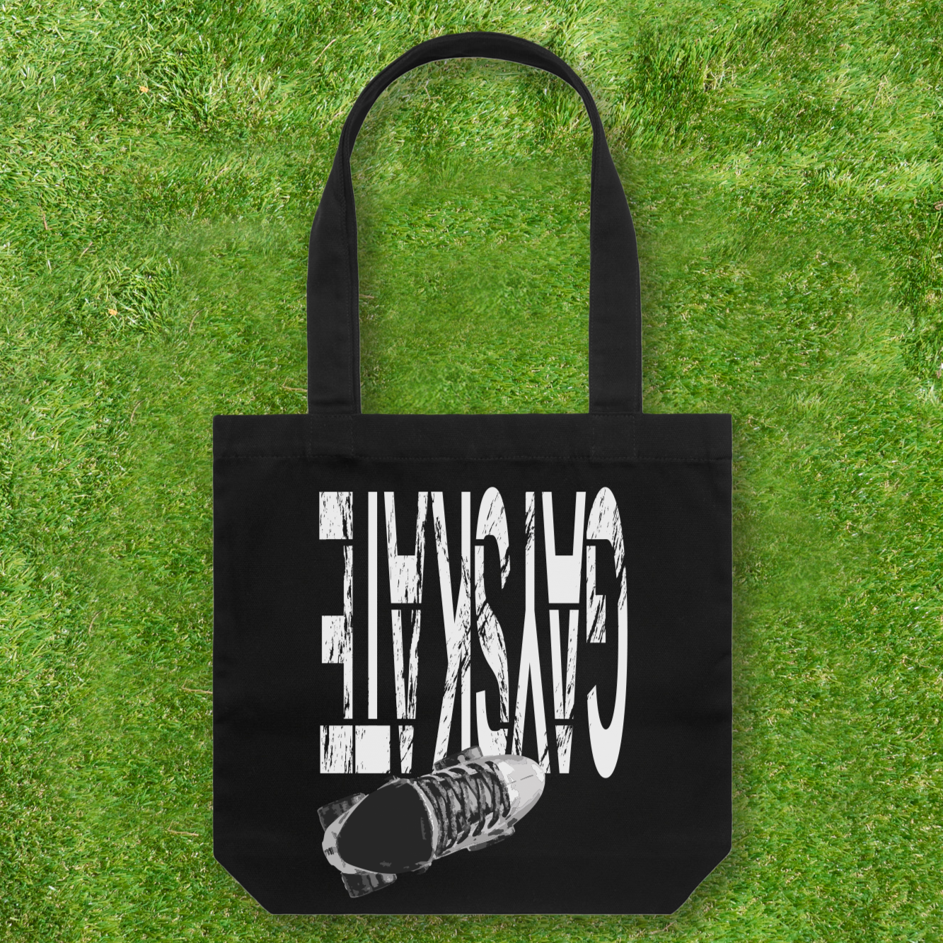 A Large printed Cotton Canvas Tote Bag - Black from Gay Skate rests on the grass, featuring "GASKATE" in white alongside an original skateboard design. Embrace style and support a queer and trans-owned business with this trendy accessory.