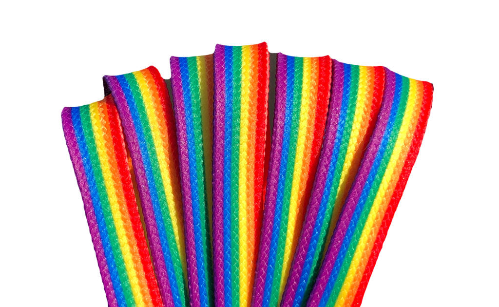 Three pairs of colorful shoelaces are displayed. The first pair is yellow with black and purple stripes, the second pair features a Pride flag pattern, and the third pair is pale pink with teal and coral stripes. All shoelaces are Waxed Skate Laces by Derby Laces, proudly presented under the Gay Skate brand.