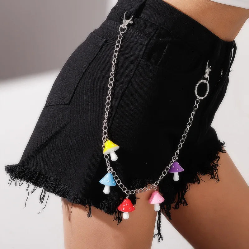 A close-up of a person wearing a denim skirt and a white belt with heart-shaped cutouts. Clipped onto the belt are three layered rainbow chains from Gay Skate's Layered Rainbow Clip-on Chains collection, boasting cute kawaii colorful designs and adorned with heart and flower charms. These chains add a playful element of vibrant cuteness to the outfit. In the background, there's greenery and a walkway.