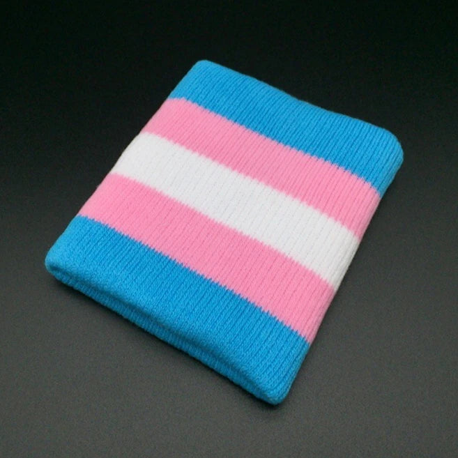 A wrist adorned with the Gay Skate Trans pride flag sporty wrist band featuring horizontal stripes in light blue, pink, and white. The hand is shown against a plain white background.