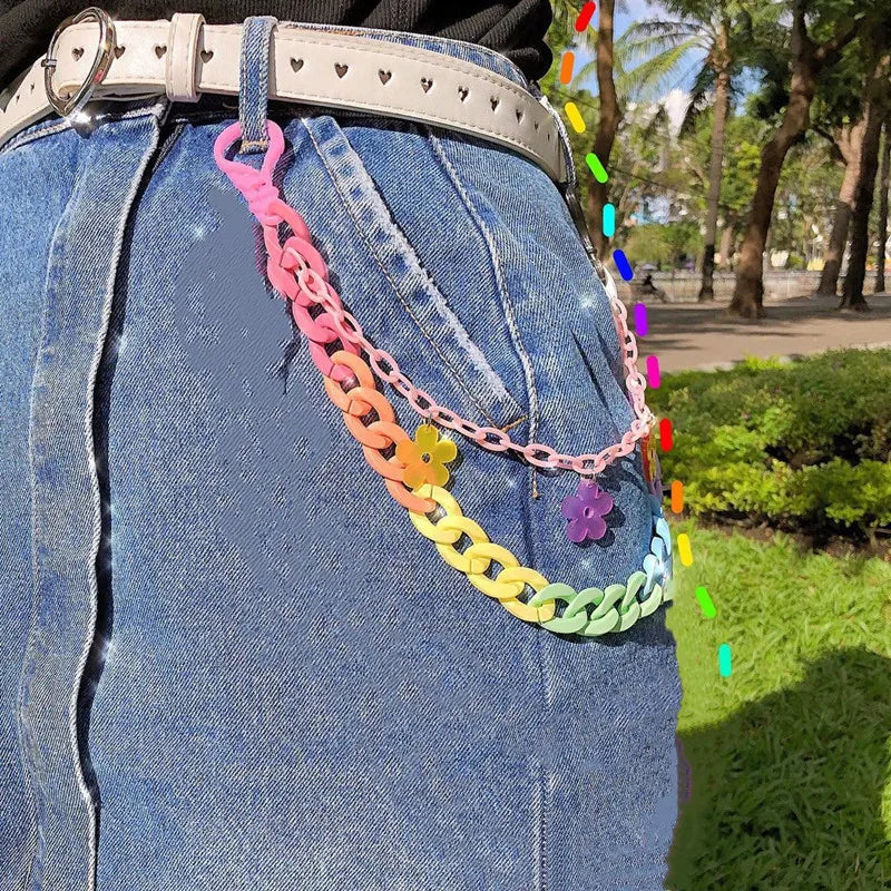 A close-up of a person wearing a denim skirt and a white belt with heart-shaped cutouts. Clipped onto the belt are three layered rainbow chains from Gay Skate's Layered Rainbow Clip-on Chains collection, boasting cute kawaii colorful designs and adorned with heart and flower charms. These chains add a playful element of vibrant cuteness to the outfit. In the background, there's greenery and a walkway.