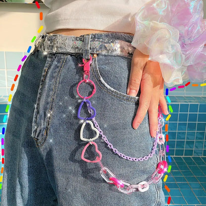 A close-up of a person wearing a denim skirt and a white belt with heart-shaped cutouts. Clipped onto the belt are three layered rainbow chains from Gay Skate's Layered Rainbow Clip-on Chains collection, boasting cute kawaii colorful designs and adorned with heart and flower charms. These chains add a playful element of vibrant cuteness to the outfit. In the background, there's greenery and a walkway.