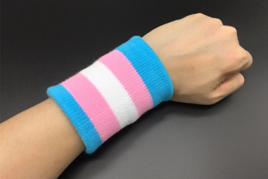 A wrist adorned with the Gay Skate Trans pride flag sporty wrist band featuring horizontal stripes in light blue, pink, and white - the transgender pride flag. An accessory for clubbing, raves, parties and festivals. Perfect for transmen, transgirls, transwomen, non-binary and other genderqueer people.