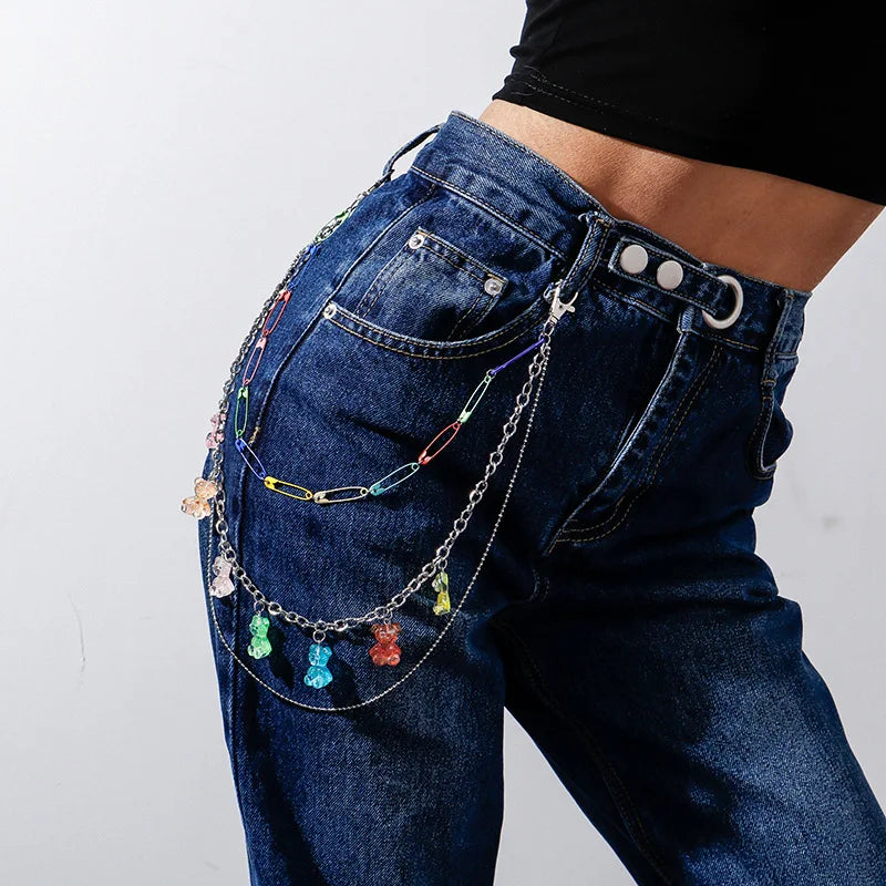 Close-up of a person wearing blue denim jeans adorned with rainbowLayered Rainbow Clip-on Chains - cute kawaii colourful designs, adding a playful harajuku element to the outfit. The chains, consisting of various colors and shapes like hearts and flowers, bring colourful cuteness. The background shows a park with green grass and trees. Acessories include carabiners, mushrooms, safety pins, gummi bears and more for punks, emos, skaters and queer fashionistas.