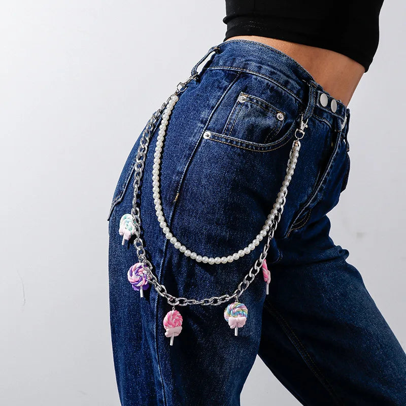 Close-up of a person wearing blue denim jeans adorned with rainbowLayered Rainbow Clip-on Chains - cute kawaii colourful designs, adding a playful harajuku element to the outfit. The chains, consisting of various colors and shapes like hearts and flowers, bring colourful cuteness. The background shows a park with green grass and trees. Acessories include carabiners, mushrooms, safety pins, gummi bears and more for punks, emos, skaters and queer fashionistas.