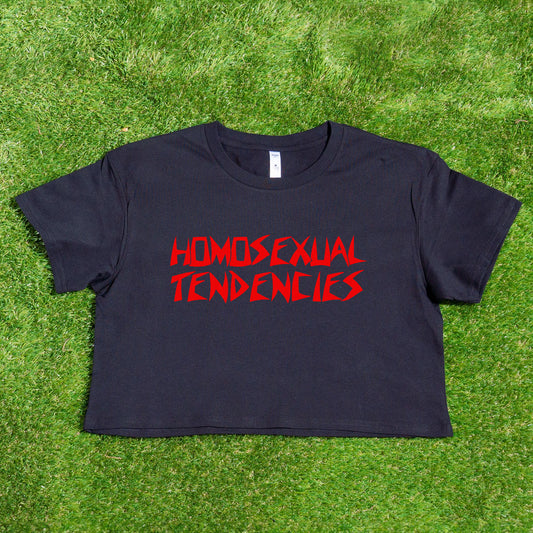 A black "HOMOSEXUAL TENDENCIES" locally printed satire crop tee from Gay Skate, styled with bold typography and celebrating queer pride, is displayed against a green grass backdrop.