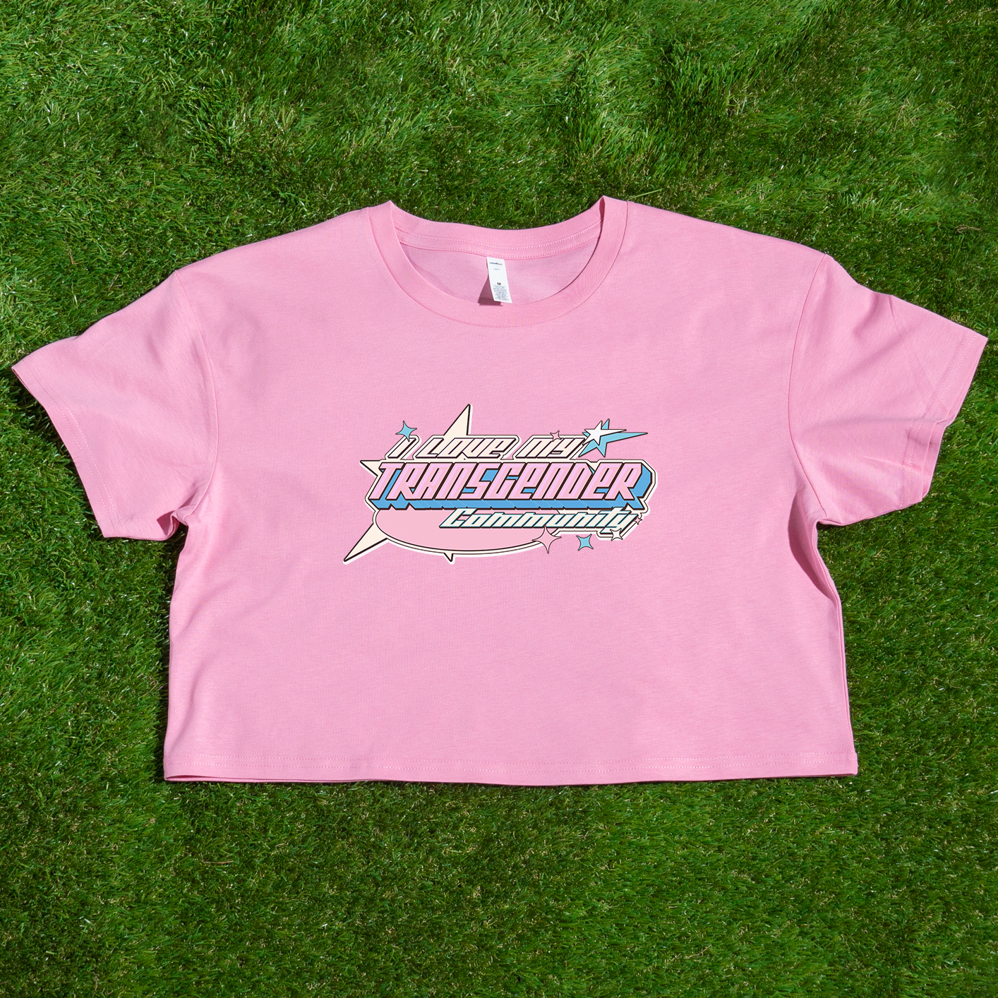 A cropped shirt from Gay Skate named "I LOVE MY TRANSGENDER COMMUNITY - cropped shirt for Australian Shipping" is laid on green grass. It features an airplane graphic, a smaller star, and bold text reading "I love my Transgender community." This pride clothing design uses the blue, white, and pink colors of the transgender pride flag.