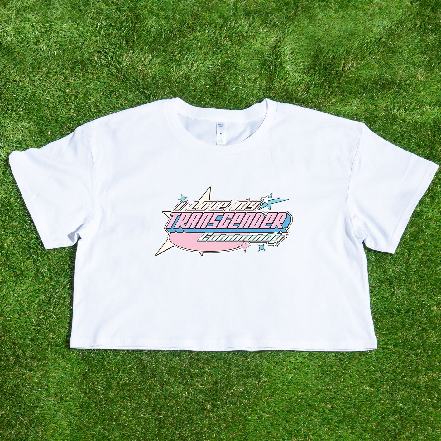 A cropped shirt from Gay Skate named "I LOVE MY TRANSGENDER COMMUNITY - cropped shirt for Australian Shipping" is laid on green grass. It features an airplane graphic, a smaller star, and bold text reading "I love my Transgender community." This pride clothing design uses the blue, white, and pink colors of the transgender pride flag.