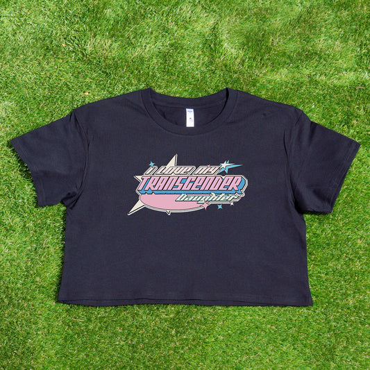 A pink crop top from Gay Skate, emblazoned with "Proud Transgender Daughter" in bold text and decorative elements, lies flat on green grass. This shirt, part of the "I LOVE MY TRANSGENDER DAUGHTER or SON" collection, boasts a classic short-sleeve design and is one of our unique original designs from a queer-owned business. Available for Australian shipping.