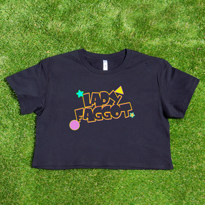 A black “Lady F@gg*t” crop tee, locally printed with colorful bold text in a playful font surrounded by stars and geometric shapes, displayed on vibrant green grass.
