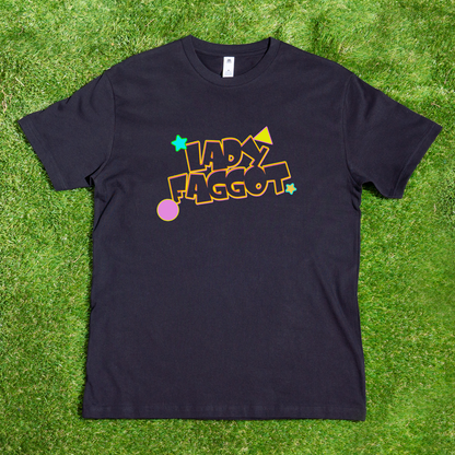 A black Lady F@gg*t - Local hand printed t-shirt lays on grass, showcasing the phrase in bold, colorful letters surrounded by stars, a circle, and geometric shapes.