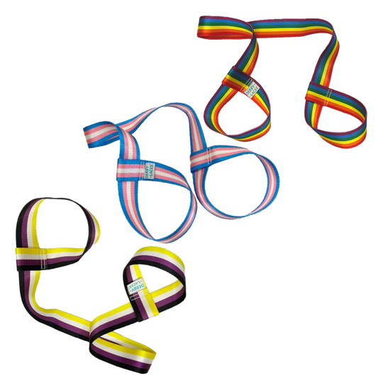 Three skate leashes are shown, each featuring unique striped color patterns: one with the rainbow flag, one with the Trans flag, and one with the Non-Binary flag. Each band has a looped design that can double as a yoga mat strap. These  skate nooses are branded under Gay Skate and named Pride Skate Leash – Gear strap.