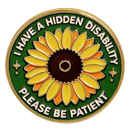 A circular enamel pin featuring a yellow sunflower on a green background with the text 'I HAVE A HIDDEN DISABILITY PLEASE BE PATIENT' in white. The pin is styled on a warm cream surface with beige satin ribbon and small white baby's breath flowers.