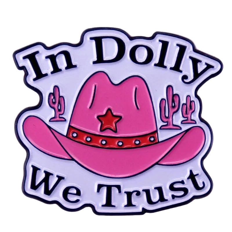 A die-cut enamel pin featuring a pink cowboy hat with a red band and star, flanked by pink cacti, with the text 'In Dolly We Trust' in black lettering on a white background with a black outline.