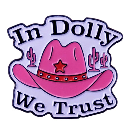 A die-cut enamel pin featuring a pink cowboy hat with a red band and star, flanked by pink cacti, with the text 'In Dolly We Trust' in black lettering on a white background with a black outline.