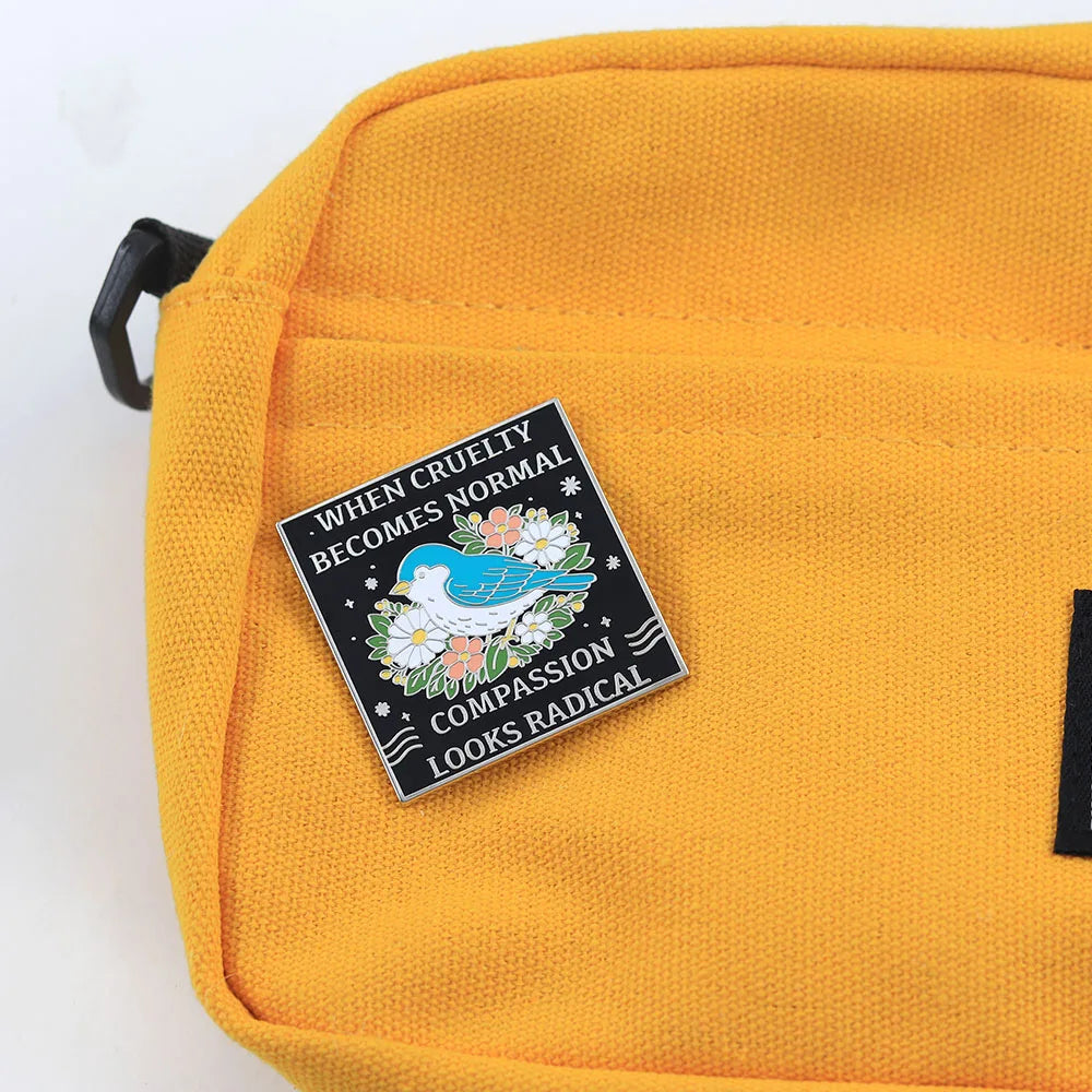 A close-up of a mustard yellow canvas bag featuring a black enamel pin with a white dove, flowers, and the text 'WHEN CRUELTY BECOMES NORMAL COMPASSION LOOKS RADICAL'.