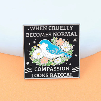 A square enamel pin with a black background featuring the text 'WHEN CRUELTY BECOMES NORMAL' and 'COMPASSION LOOKS RADICAL' in white lettering, surrounding an illustration of a blue bird with white, pink, and green flowers.