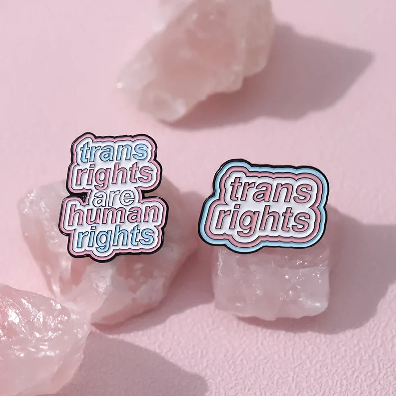 Product Description: The "Trans Rights and Trans Pride" enamel pins collection by Gay Skate includes six beautifully designed badges celebrating transgender pride. Top row designs feature: "Trans Rights Are Human Rights," "Trans Rights," and "Respect Trans Women Juice." Bottom row designs include an anatomical heart with trans flag colors, a heart with layered trans flag colors, and a rainbow with trans flag colors.