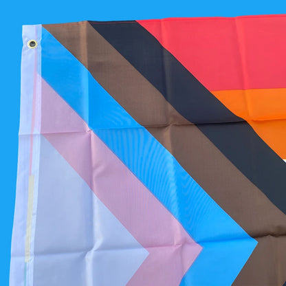 This image showcases the 150cm x 90cm Progress Pride Rainbow Flag by Gay Skate. It is made of high-quality polyester and features traditional rainbow stripes, along with a chevron on the left that includes black and brown stripes, plus light blue, white, and pink stripes to represent marginalized LGBTQ+ communities, including people of color and the trans community.