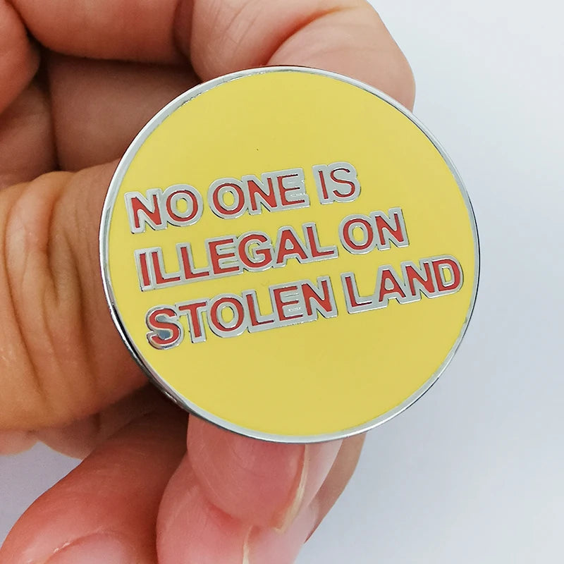 A hand holding a round yellow enamel pin with the text 'NO ONE IS ILLEGAL ON STOLEN LAND' in red letters with white outlines, set against a plain white background.