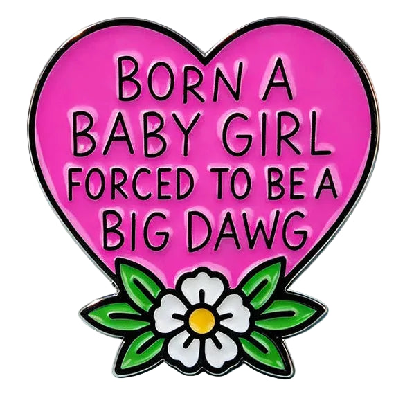 A heart-shaped enamel pin with the text 'BORN A BABY GIRL FORCED TO BE A BIG Dawg' in black on a bright pink background, decorated with green leaves and a white flower with a yellow center at the bottom. 