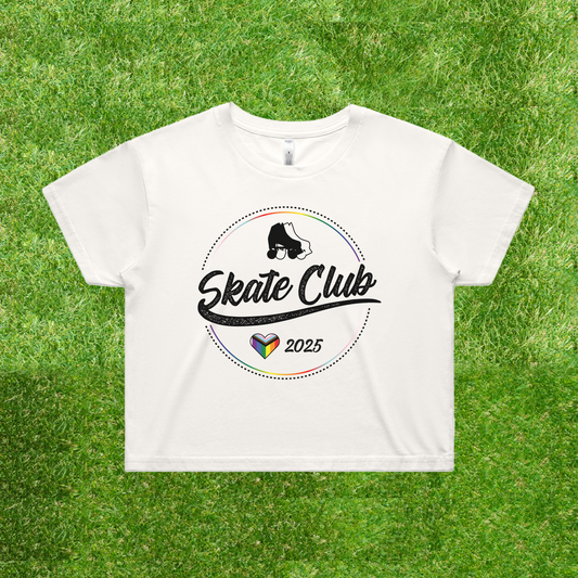 A white crop tee features the "SKATE CLUB 2025" design with a skateboarder's silhouette and a rainbow-colored heart, echoing the progress pride flag. Set against a grassy background, this locally printed shirt embodies the Gay Skate brand.