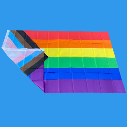 This image showcases the 150cm x 90cm Progress Pride Rainbow Flag by Gay Skate. It is made of high-quality polyester and features traditional rainbow stripes, along with a chevron on the left that includes black and brown stripes, plus light blue, white, and pink stripes to represent marginalized LGBTQ+ communities, including people of color and the trans community.