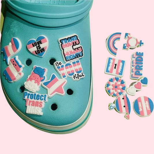 The "Trans Pride Shoe Charms/Pins/Badges" from Gay Skate are blue shoes decorated with 16 vibrant pins that prominently feature designs and text supporting trans rights. These include slogans such as "Love is Love" and "Protect Trans Kids," accompanied by pride flags, hearts, and rainbow motifs, all celebrating transgender identity on a light pink background.