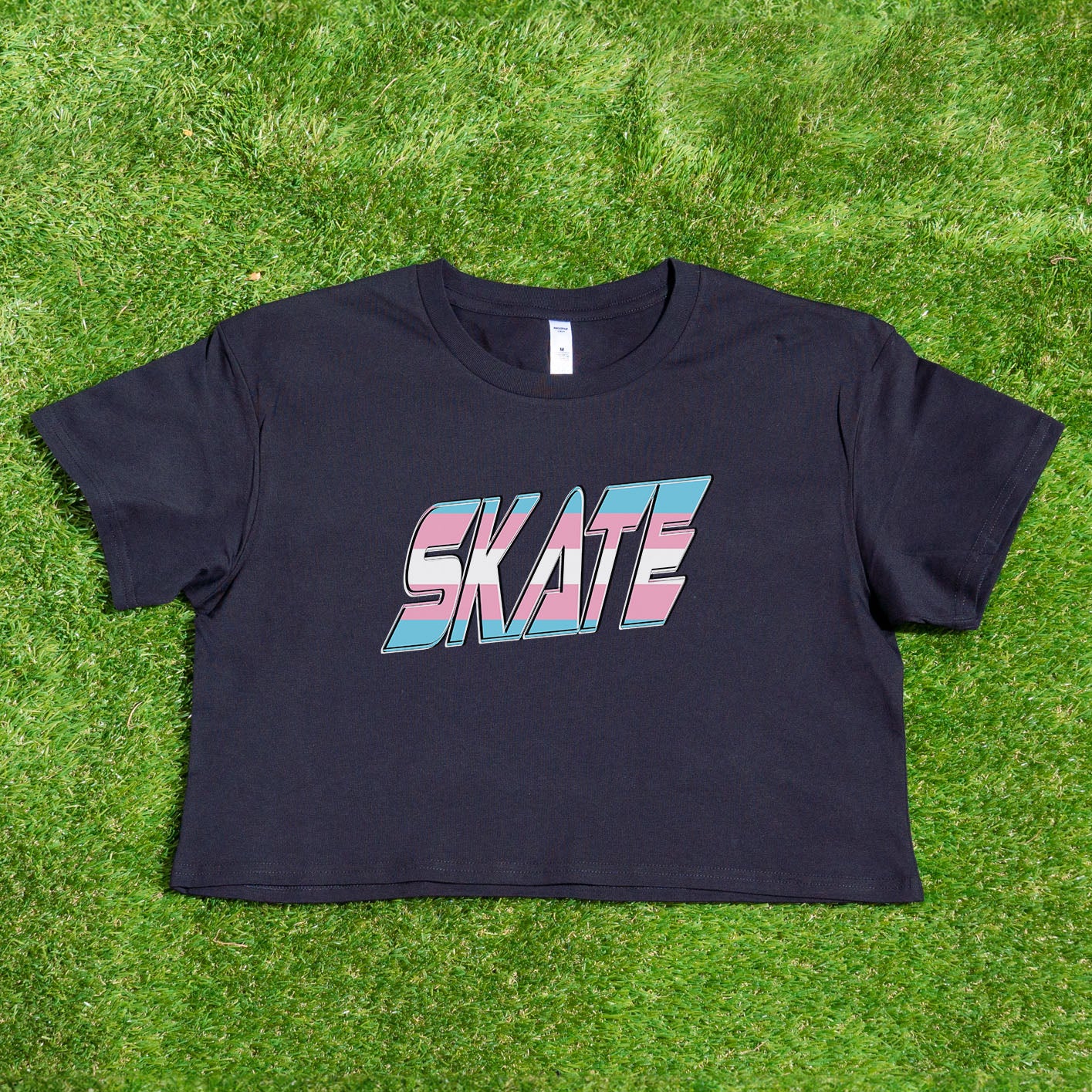 The "SKATE Trans Flag - locally printed crop tee" by Gay Skate is a black T-shirt featuring the word "SKATE" in bold, colorful, and stylized letters on the front. The letters have a gradient effect with shades of pink, blue, and white reminiscent of the transgender flag, giving the design a vibrant and dynamic appearance. The T-shirt has a cropped fit perfect for streetwear fashion.