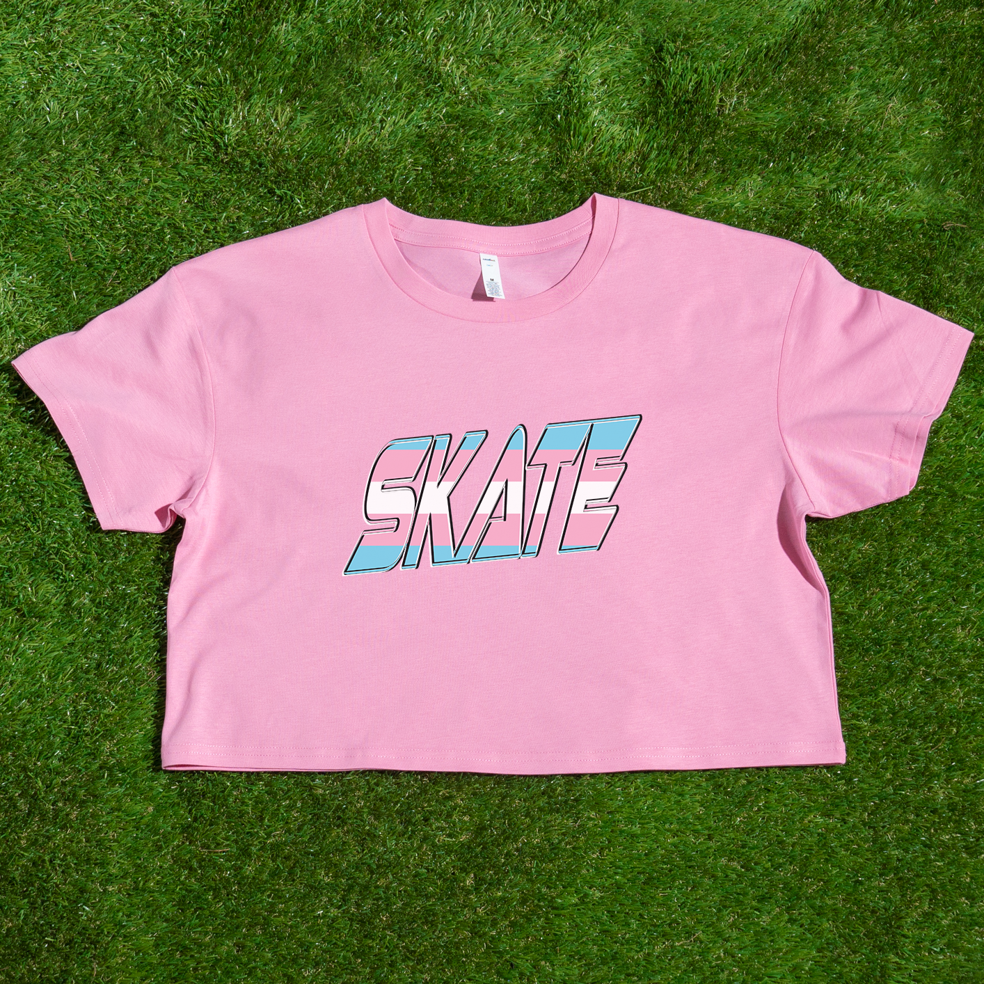 The "SKATE Trans Flag - locally printed crop tee" by Gay Skate is a black T-shirt featuring the word "SKATE" in bold, colorful, and stylized letters on the front. The letters have a gradient effect with shades of pink, blue, and white reminiscent of the transgender flag, giving the design a vibrant and dynamic appearance. The T-shirt has a cropped fit perfect for streetwear fashion.