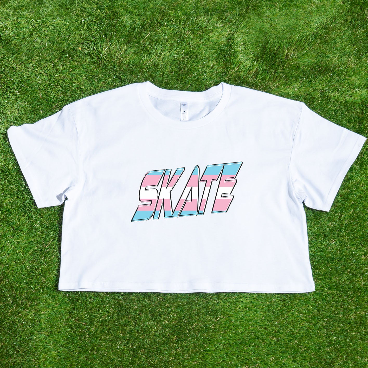 The "SKATE Trans Flag - locally printed crop tee" by Gay Skate is a black T-shirt featuring the word "SKATE" in bold, colorful, and stylized letters on the front. The letters have a gradient effect with shades of pink, blue, and white reminiscent of the transgender flag, giving the design a vibrant and dynamic appearance. The T-shirt has a cropped fit perfect for streetwear fashion.