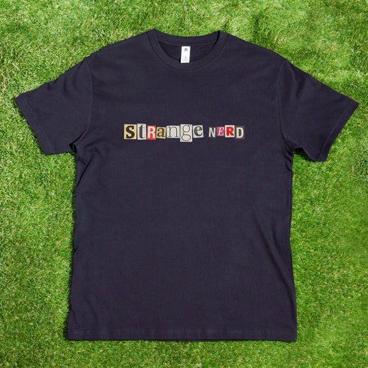 A black Gay Skate T-shirt lies on the grass, proudly displaying "Strange Nerd Transgender Anagram" in multicolored, ransom-note style letters across the front.