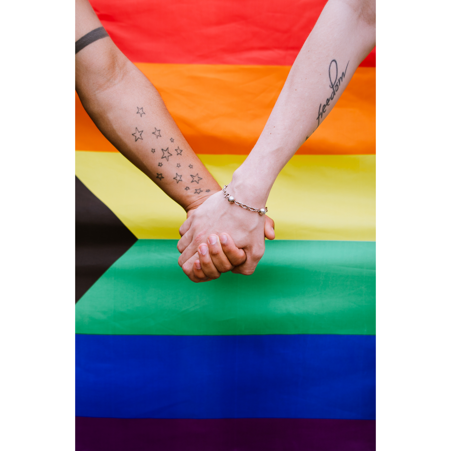 Two hands holding each other in front of a large 3ft x 5ft Gay Skate "Large Pride Flag: Progress Pride Rainbow, Trans, Non-binary, Lesbian, Pan and many more." One arm has tattoos of stars, and the other has the word "freedom" tattooed on it. The background features the inclusive pride flag with additional black, brown, light blue, pink, and white stripes made from durable polyester.