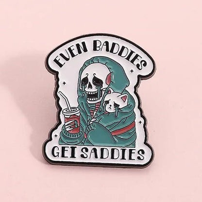 Enamel pin with a skeleton holding a cat and text 'Even Baddies Get Saddies' 