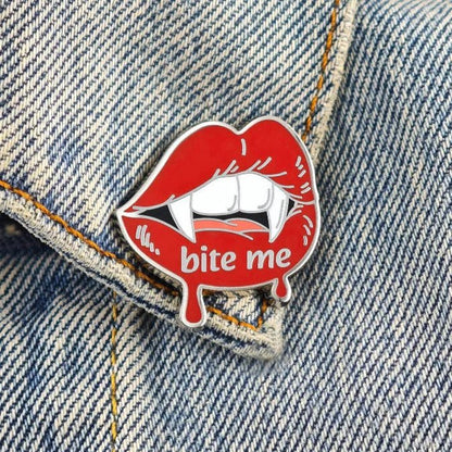 A red enamel pin shaped like lips with white fangs and the text 'bite me' attached to a blue denim jacket.