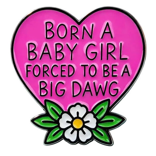A heart-shaped enamel pin with the text 'BORN A BABY GIRL FORCED TO BE A BIG Dawg' in black on a bright pink background, decorated with green leaves and a white flower with a yellow center at the bottom. 