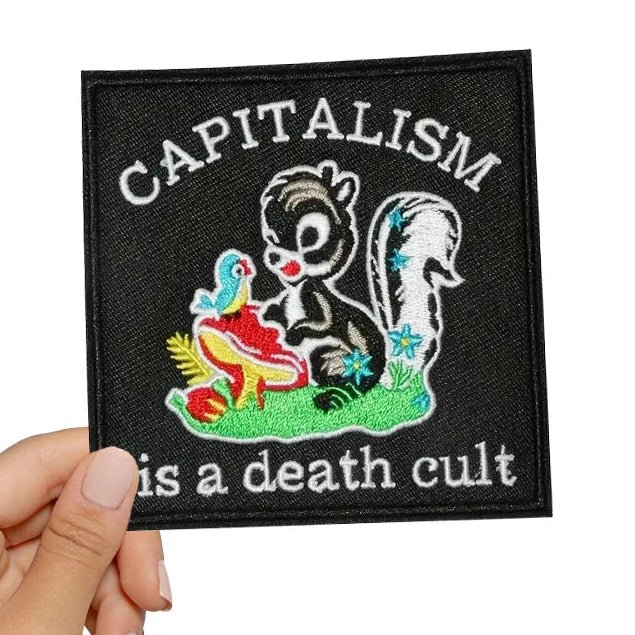 Patch with a cartoon squirrel and bird, text 'CAPITALISM is a death cult', held by a hand on a white background