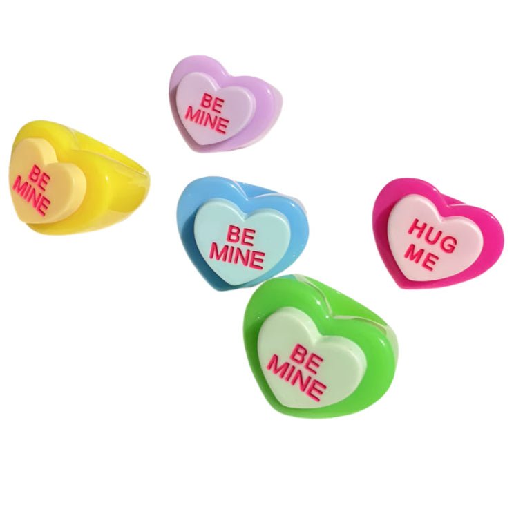 Gay Skate's Chunky Resin candy heart rings feature colorful acrylic hearts with phrases "BE MINE" and "HUG ME" in red. Available in pink, blue, green, yellow, and purple, each design features one heart playfully overlapping another.