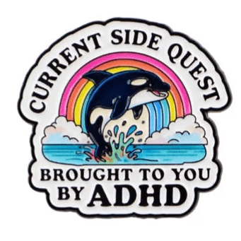 The "Current Side Quest Brought To You By ADHD" enamel pin features a cartoon orca leaping from the ocean in front of a rainbow, with the phrase encircling the colorful scene.